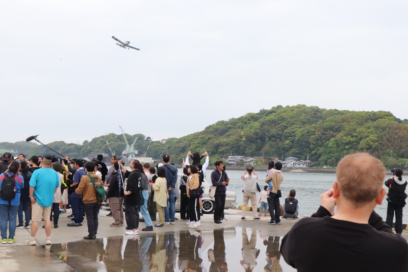 アクロバット飛行を行う飛行機を見上げ、カメラを向ける来場者の写真