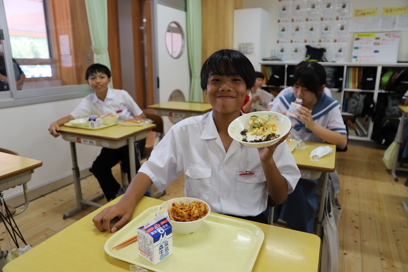 皿に入った給食の石炭ナッツピーを手に取り、笑顔の生徒と給食を食べている生徒の写真